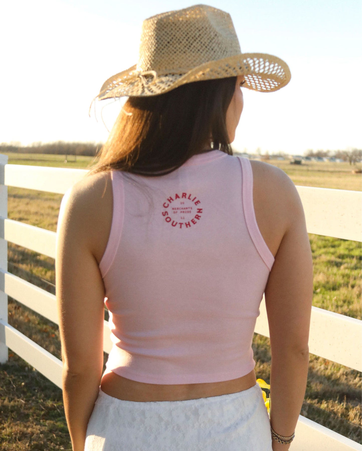 Back view of a woman wearing a Women's Rodeo Queen Tank Top by Charlie Southern, showcasing the brand logo on the back while standing outdoors near a white fence.