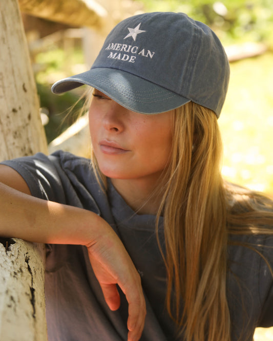 Wearing a Women's American Made Cap by Charlie Southern, featuring a clean white embroidered star on washed denim. The model enjoys a sunny outdoor setting, showcasing the cap's casual style.