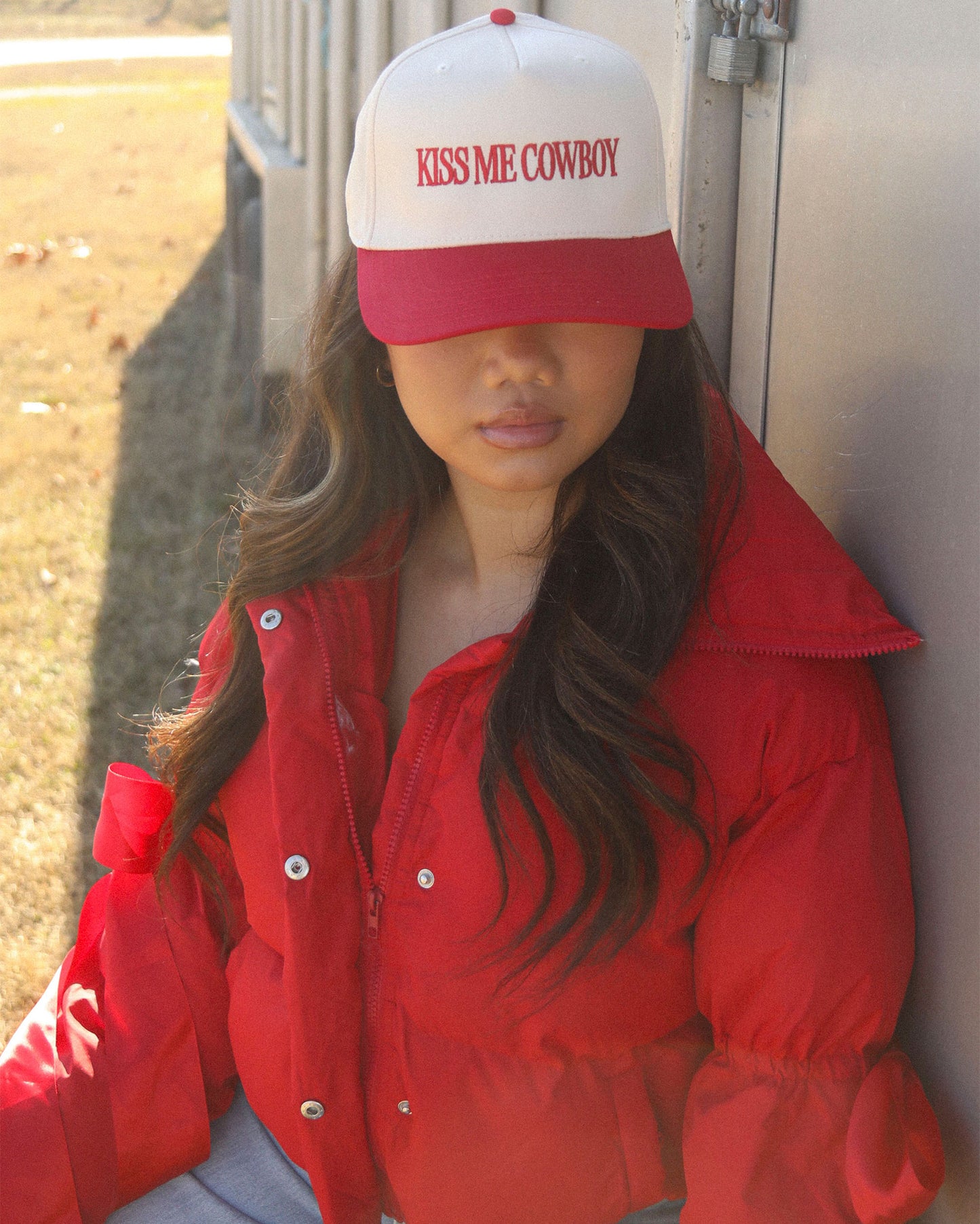 A stylish woman wearing the Women's Kiss Me Cowboy Trucker Cap by Charlie Southern, featuring a white front and red brim. The cap showcases the phrase 'KISS ME COWBOY' in bold lettering, paired with a cozy red puffer jacket, set against a sunny outdoor background.