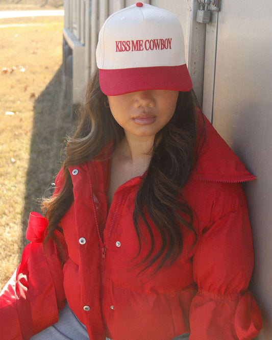 A stylish woman wearing the Women's Kiss Me Cowboy Trucker Cap by Charlie Southern, featuring a white front and red brim. The cap showcases the phrase 'KISS ME COWBOY' in bold lettering, paired with a cozy red puffer jacket, set against a sunny outdoor background.